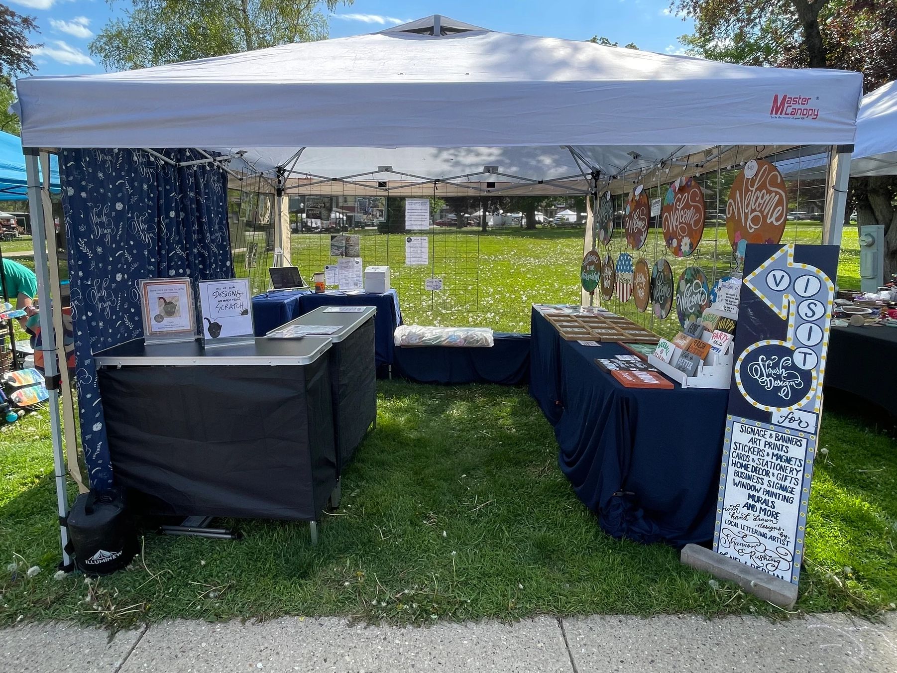 A neatly arranged outdoor art and signage booth under a white canopy tent.