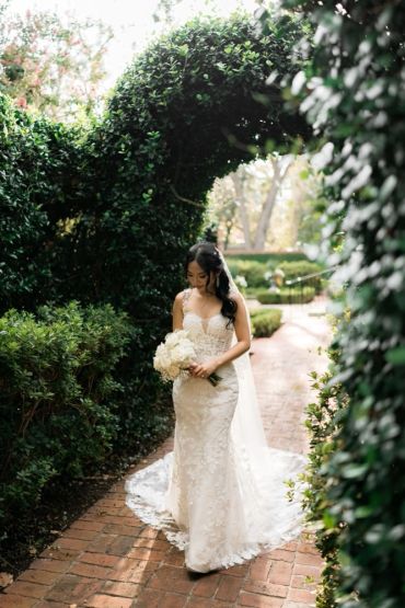 Bride in a lace wedding gown holding a bouquet under a green archway.