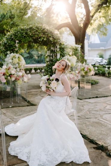 Bride in white gown holding bouquet in sunlit garden.