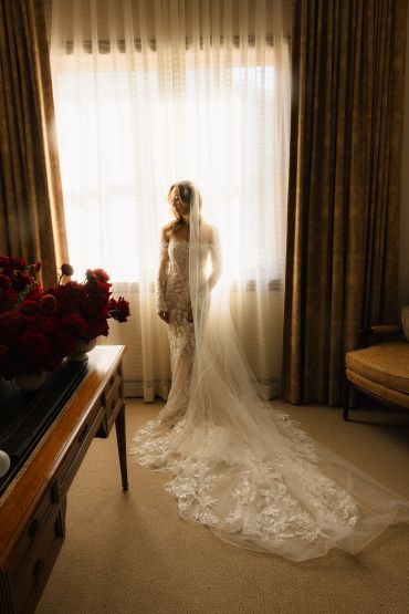 Bride in lace gown and veil stands by a sunlit window.