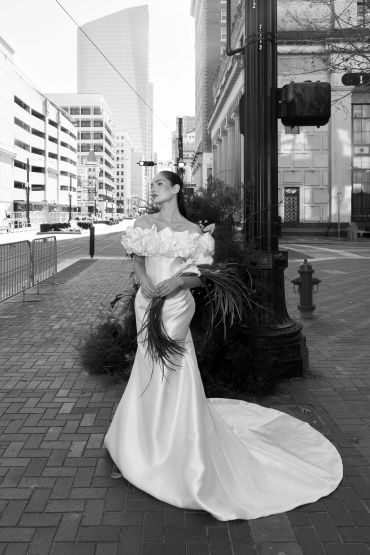 Elegant bride in a flowing gown holds greenery on a city street.