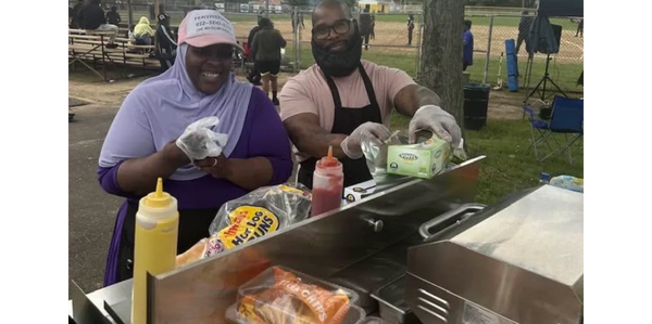 Two people happily preparing food at an outdoor event with hot dog buns and condiments.