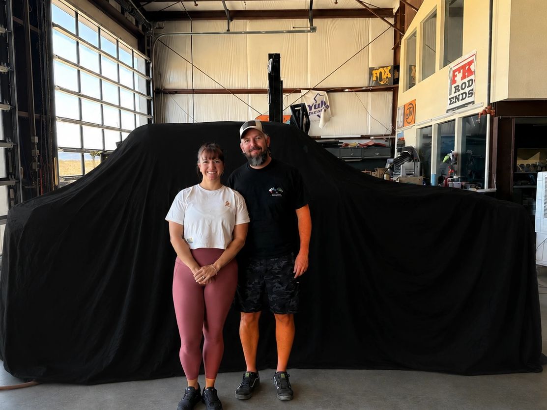 A smiling couple stands in front of a covered vehicle in a garage.