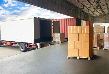 Rear of a white delivery truck backed up to a warehouse, with stacks of boxes on pallets.





