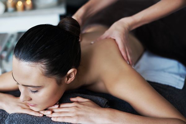 A woman with her hair in a bun is lying on her stomach, receiving a back massage from another person