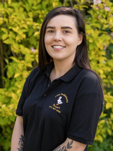 Smiling woman in a black nursery polo shirt stands outdoors against green foliage.