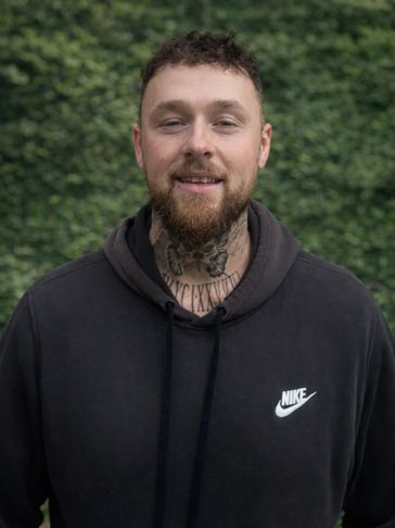 Smiling man with curly hair and neck tattoo wearing a black Nike hoodie.