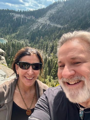 Smiling couple taking a selfie with forested mountains in the background.