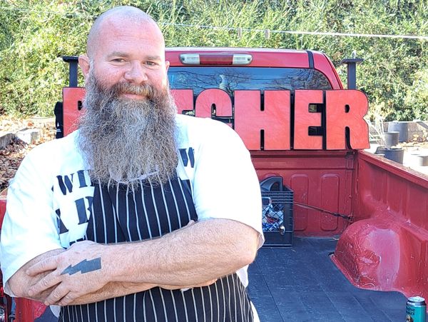 Bearded man in apron standing confidently by a truck bed with large red letters.