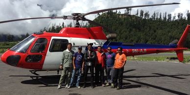 Six men stand in front of a red and white helicopter on a sunny day.