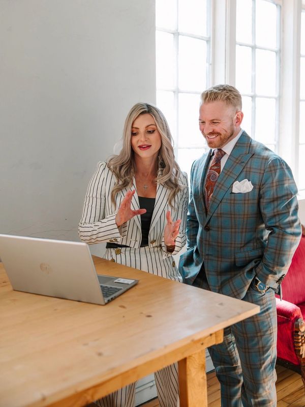 Two professionals in suits discussing work by a laptop in a bright room.