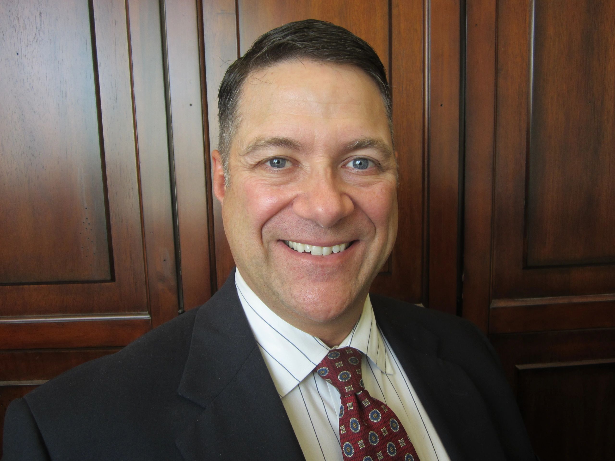 Smiling man in business suit and patterned tie standing before wooden panels.
