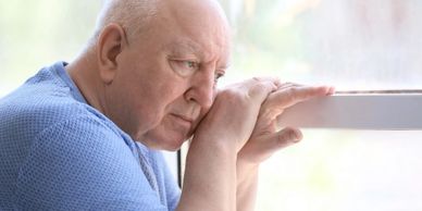 Elderly man looking thoughtfully out a window with a somber expression.
