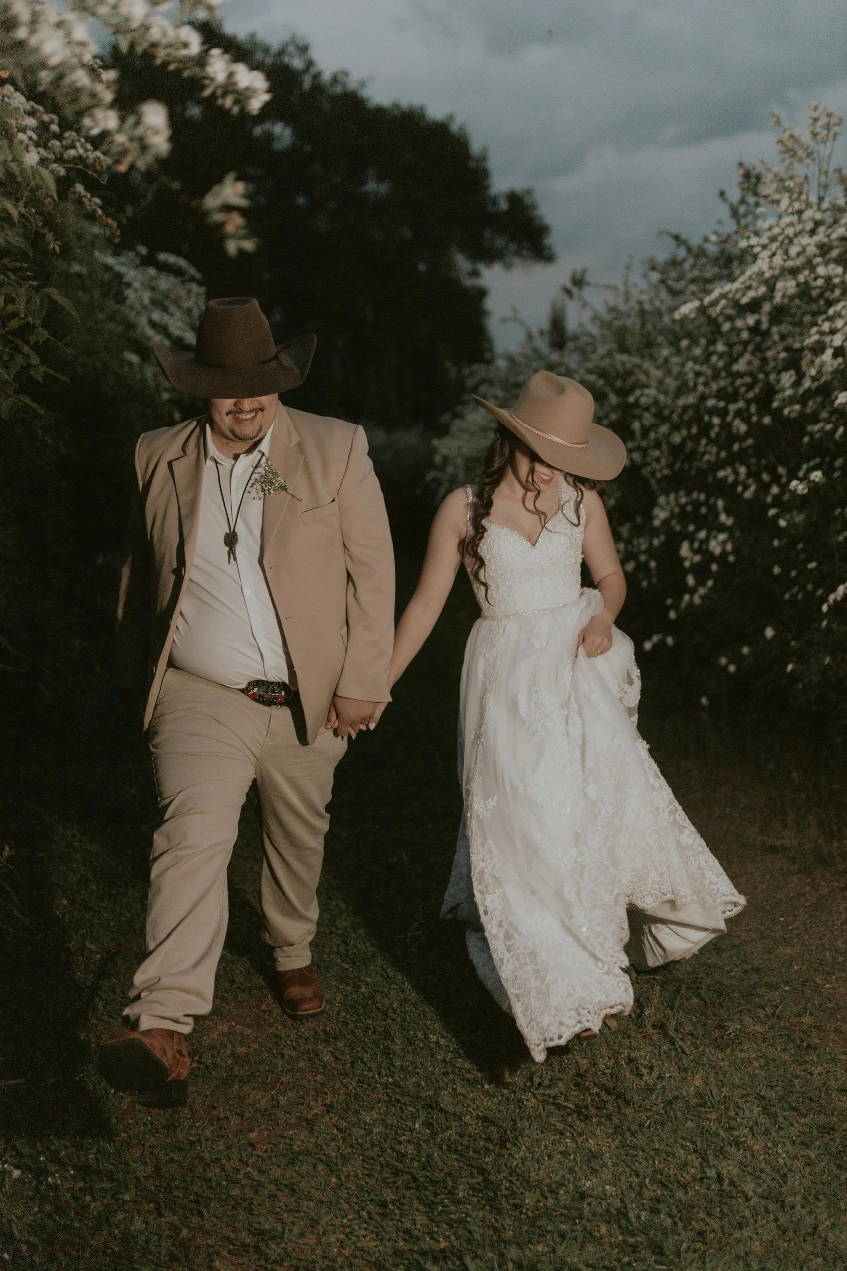 A couple in wedding attire walking hand in hand wearing cowboy hats.