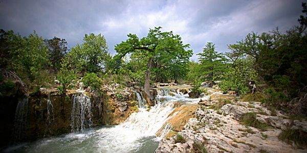 A scenic waterfall flowing over rocky terrain surrounded by green trees under a cloudy sky.