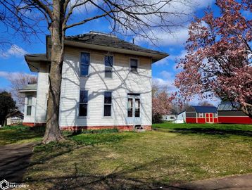 Two-story white house with blossoming trees and red sheds under a blue sky.