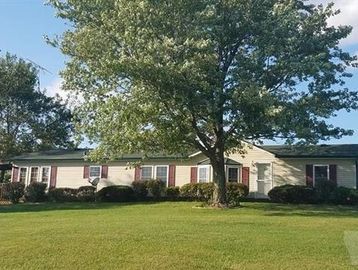 A single-story house with beige siding and maroon shutters behind a large tree on a grassy lawn.