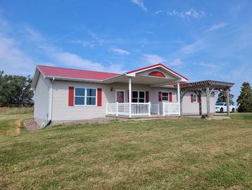 Single-story house with red roof, red shutters, and a wooden pergola on a grassy lawn.