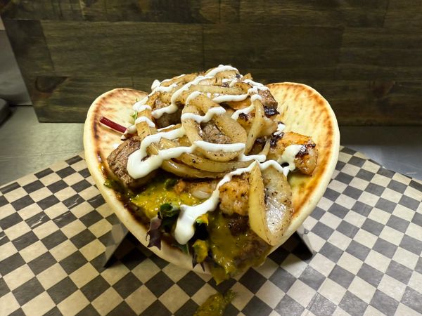 A flatbread sandwich with grilled meat, onions, greens, and sauce on checkered paper.