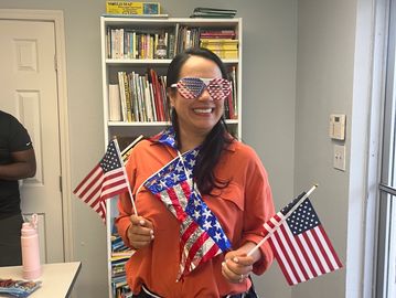 A woman wearing star-spangled glasses holds three American flags indoors.