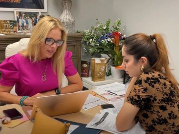 Two women discussing work at an office desk with documents and a laptop.