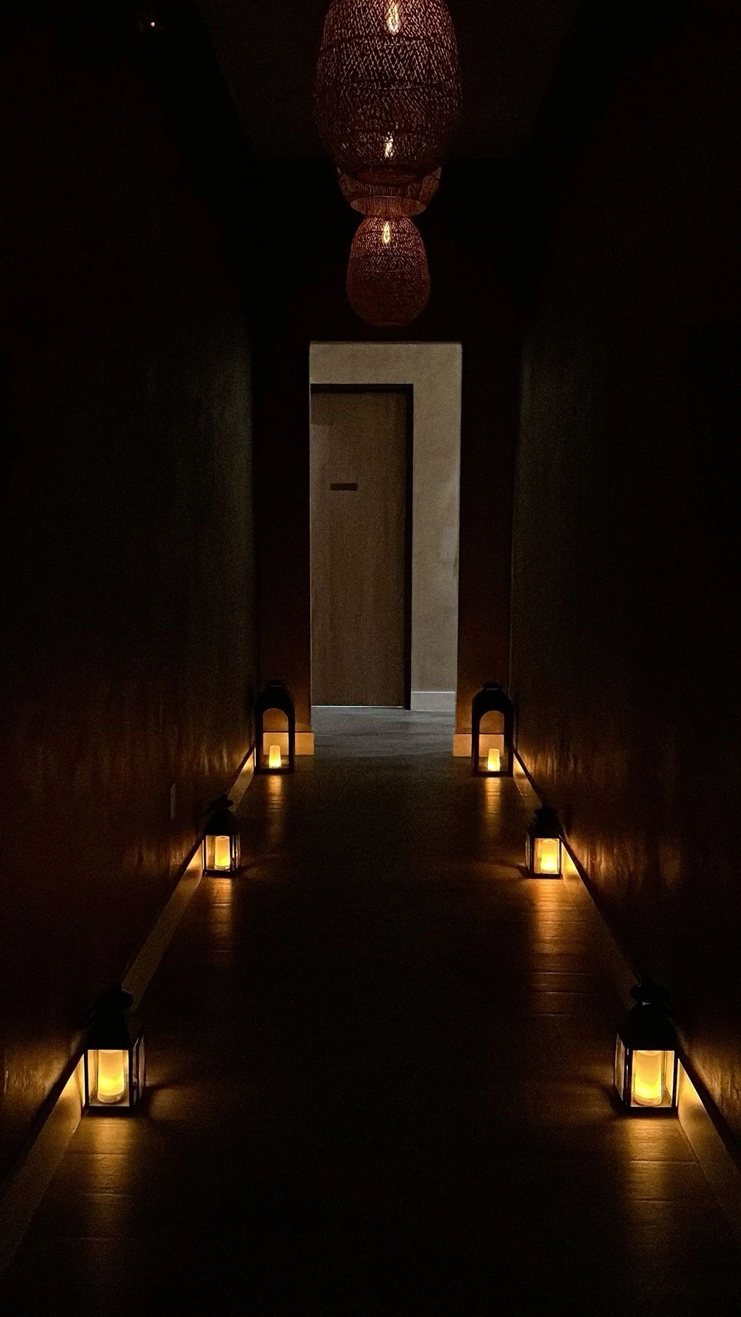 Dimly lit corridor with lanterns and hanging lamps leading to a closed door.