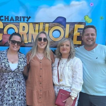 Four people posing in front of a charity cornhole sign.