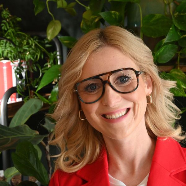 Smiling woman with glasses and blonde hair in front of plants.