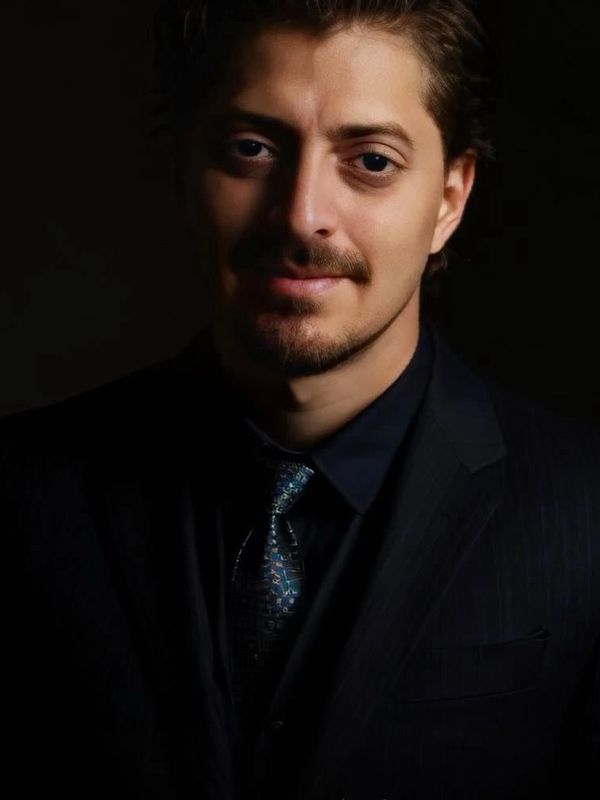 Portrait of a man in a dark suit and patterned tie against a black background.