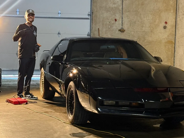 Man posing next to a black sports car in a garage.