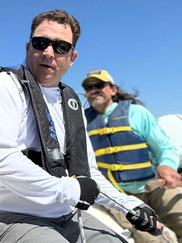 Two men wearing life jackets sailing on a boat under a clear blue sky.