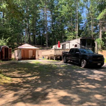 Black pickup truck parked near a mobile home and tents in a wooded area.