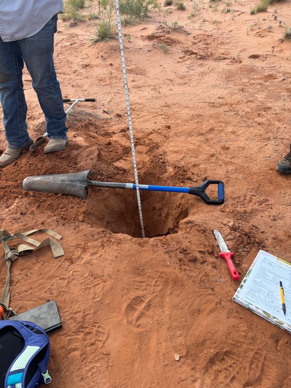 Shovel resting over a deep hole with a measuring stick inside on red soil.