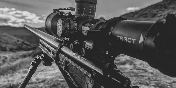 Close-up of a sniper rifle scope set against an outdoor landscape.