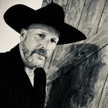 Man with beard wearing a wide-brimmed hat in front of wooden door.