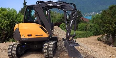 A yellow Mecalac excavator digging soil on a construction site with mountains in the background.