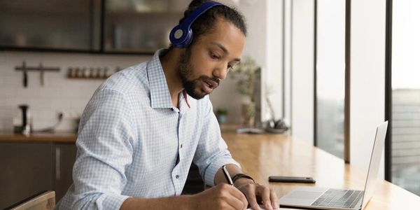 Man wearing headphones, writing notes while working on laptop.