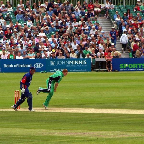 Cricket bowler delivering ball with batsman ready to play in front of a large crowd.