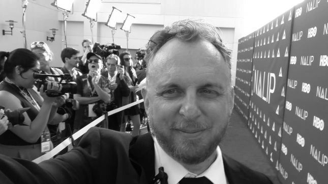 Man in suit taking selfie at a media event with photographers in background.