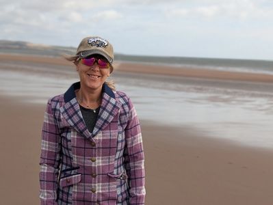 Woman in a plaid jacket and cap standing on a beach with a calm sea in the background.