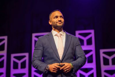 A confident man in a suit stands on stage with purple lighting.