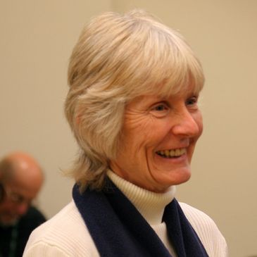 Smiling elderly woman with gray hair in a white turtleneck and dark scarf.