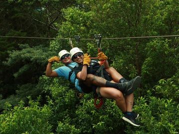 Two people zip-lining through lush green forest wearing helmets and gloves.