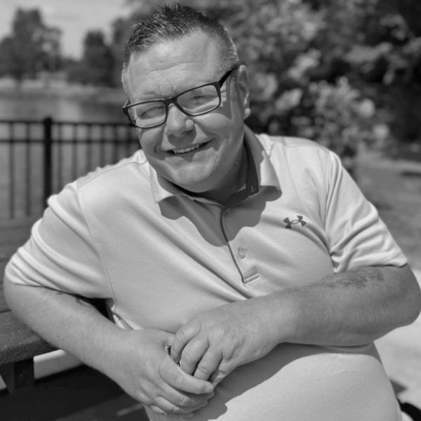 Man in blue polo shirt smiling outdoors near a lake.