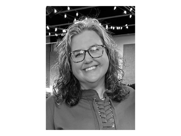 Smiling woman with glasses and curly hair wearing a brown lace-up top.