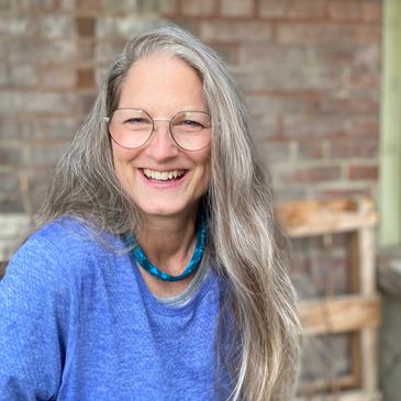 Smiling woman with long gray hair and glasses in a blue sweater.