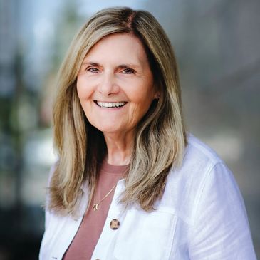 Smiling older woman with blonde hair wearing a white jacket and brown top.