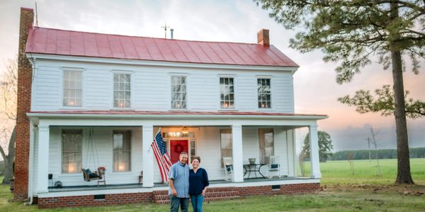 Couple standing in front of a classic white farmhouse with an American flag.
