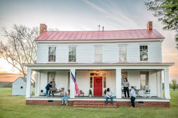 Family enjoying time on the porch of a traditional white farmhouse at sunset.