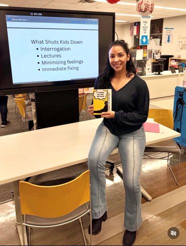 Woman holding a book, sitting on a table in a classroom with a presentation slide behind her.